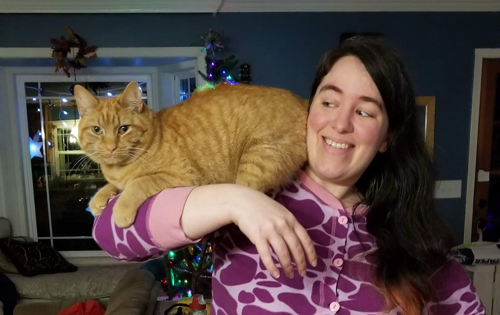 A photo of the author wearing purple cow-spot footie pajamas with a very happy orange cat named Thomas TumTum McRubbins perched on her shoulder and lifted right arm.