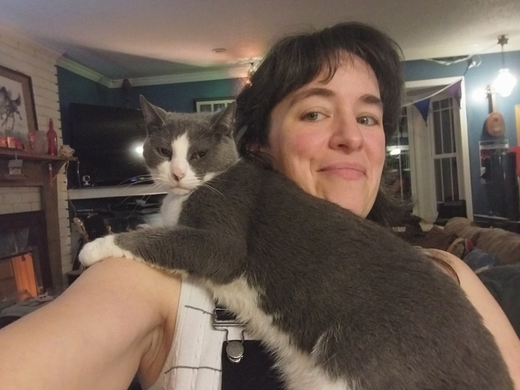 A grey and white cat draped over Lucy's shoulder, glaring at the camera while Lucy stifles a smile, taking a selfie.