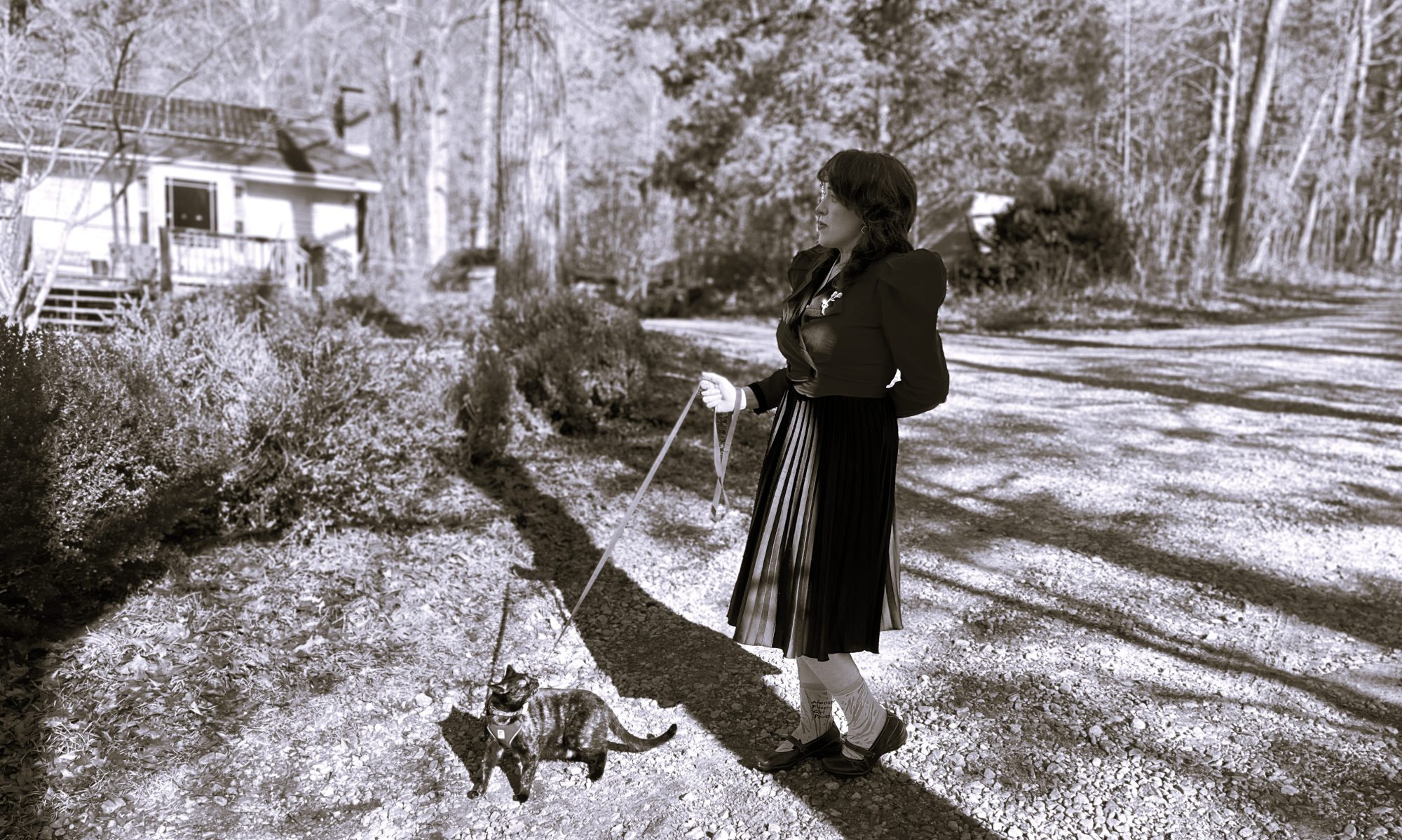 A black and white photo of Lucy K.R. in a period jacket with puffy sleeves, regarding the skyline while taking a tortoieshell cat out for a walk on a leash.