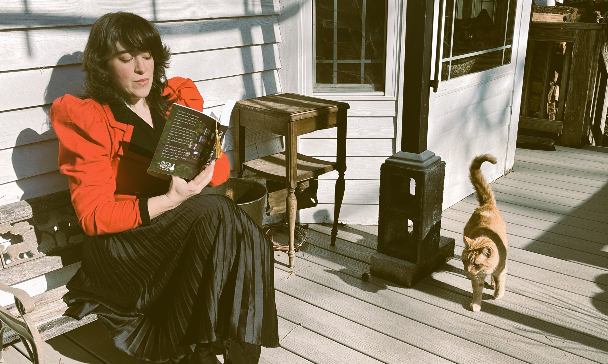Lucy K.R. wearing a period red jacket with puffy sleeves, reading on a porch while being approached by a friendly orange cat.