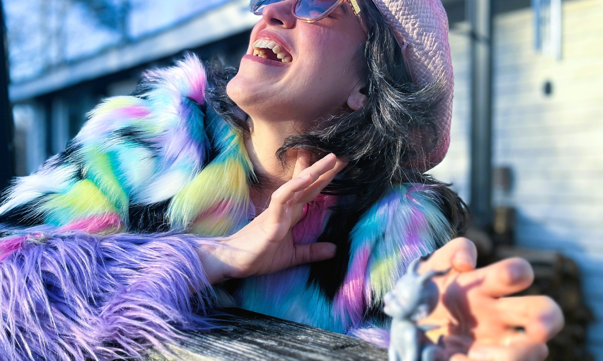 Lucy in a pink cowboy hat with sunglasses and a multicolored furry jacket that looks like it's made of furbies laughs wildly while cradling a small statue of a demon child.