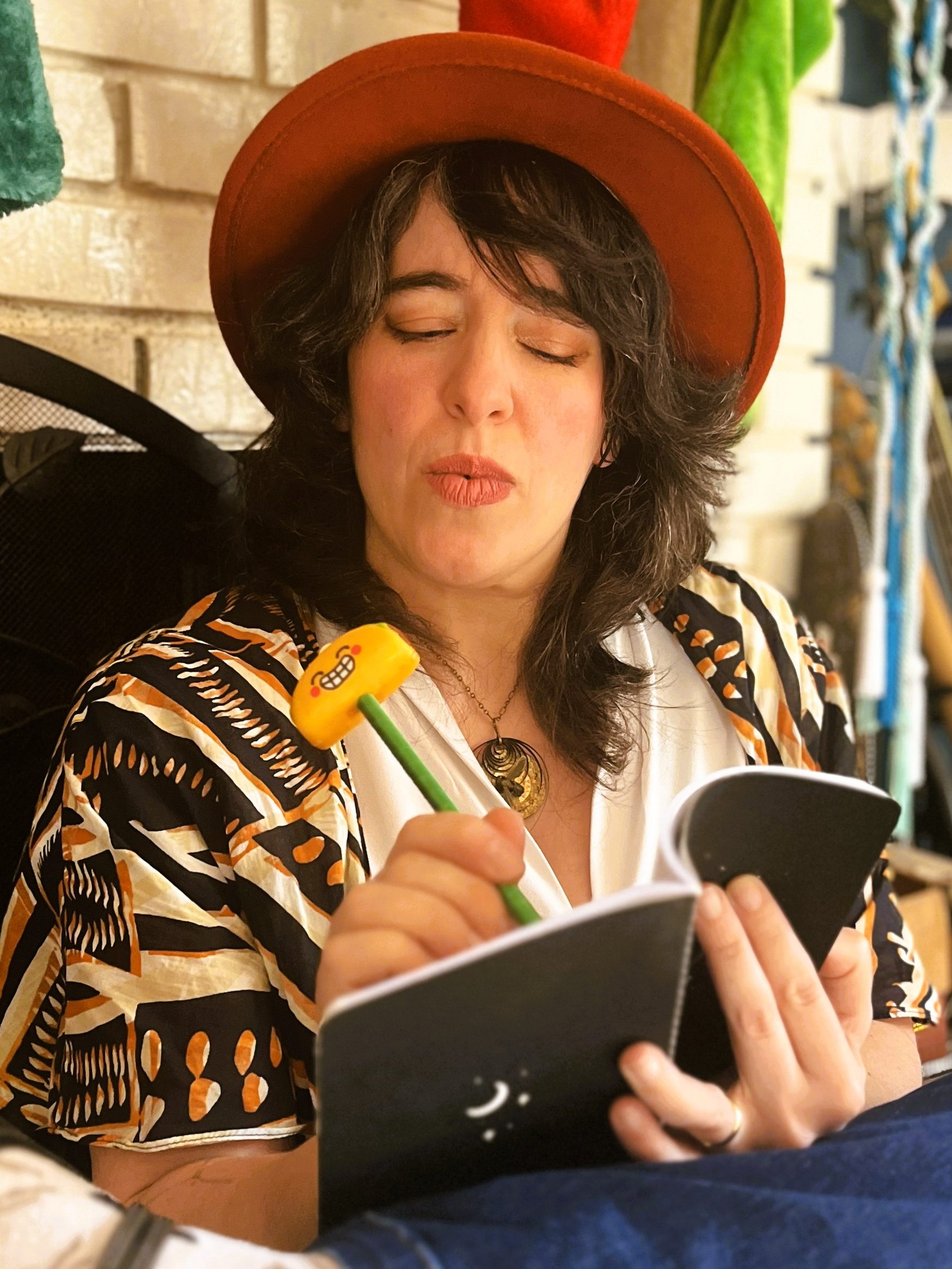 Lucy K.R. pretends to write seriously in a black notebook, legs performatively crossed, using a silly pen with a grinning taco for an eraser.