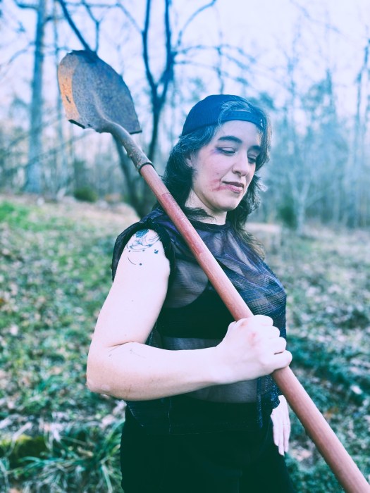 Lucy holding a shovel over her shoulder, with smudged makeup, butch sleeveless iridescent top, and a backwards baseball cap.